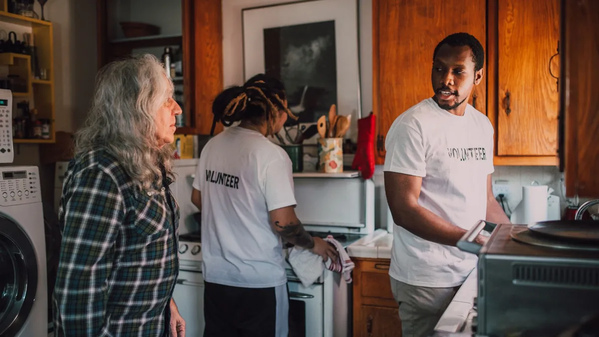 Volunteers interact and assist a senior in a cozy kitchen environment.