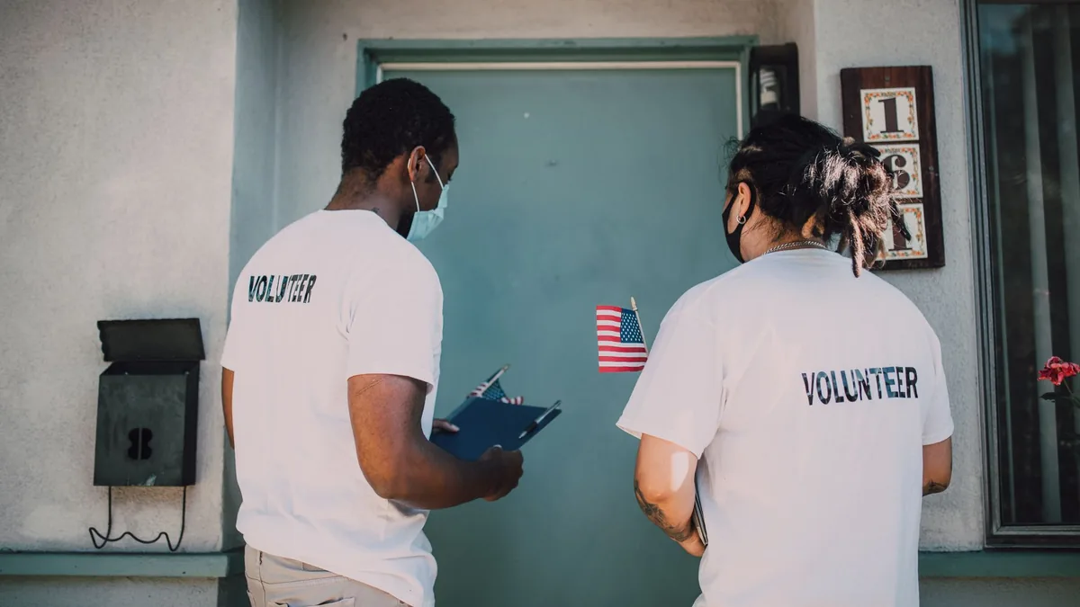 Two volunteers wear masks and visit community homes, engaging in outreach activities.